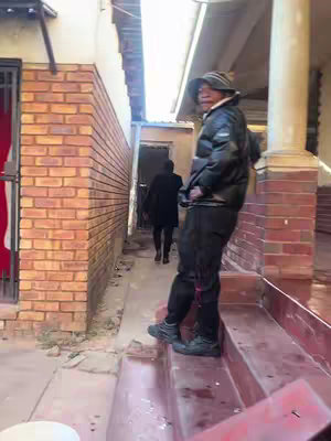 A man stands on a step, looking towards the camera and slightly to the right. Another person in dark clothing walks down the narrow passage between two buildings. The image is taken from near a front door or entrance; bricks can be seen on a building to the left, and red steps are in the foreground.