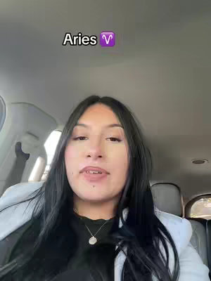 A woman with long black hair sits in a car, facing the camera. She's wearing a black top under a light-colored jacket and a silver necklace. Above her head is a text overlay that says 'Aries' with the Aries symbol.