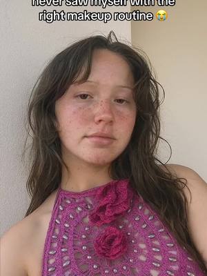 A selfie-style shot of a young woman with long brown hair and freckles. She is wearing a pink, intricately designed top with a halter neckline and two decorative rose-like embellishments. The background is a blurred, neutral color, likely a wall. The woman has a slightly introspective expression.