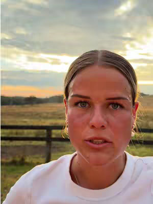 A close-up shot of a woman's face against an outdoor late afternoon or early evening backdrop. The woman has fair skin, light eyes, and her hair is pulled back. She has a neutral expression with a slight focus, likely speaking. The background suggests an open field with trees, a fence, and a sky featuring a gradient of warm colors.