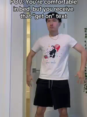 A young man stands in his home, caught mid-action, framed in a wide shot against a light-colored doorframe and wall. He's wearing a white t-shirt with a print, black shorts, and has a casual expression. Text overlays are placed at the top.