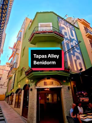 An eye-level shot of a narrow alleyway in Benidorm old town. The buildings are tall, with green walls and balconies. A sign for "GRUPO AURRERA" is visible on the side of the building on the right. In the foreground, the entrance to a restaurant, BODEGON, is under a small sign.