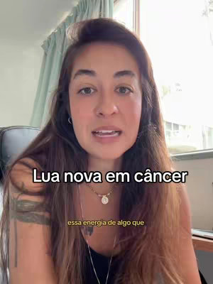 A woman with long brown hair is centered in the frame, speaking to the camera against a soft-focus background of a curtain and a window. Text reading 'Lua nova em câncer' is overlaid above her, and additional text below. The composition is a medium shot, focusing on her face and upper body.