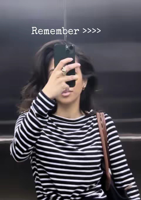 A selfie-style shot taken in an elevator, featuring a woman holding a phone up to her face, partially obscuring it. She's wearing a black and white striped long-sleeve shirt, with her dark hair around her face. The text 'Remember >>>>' is overlaid near the top.