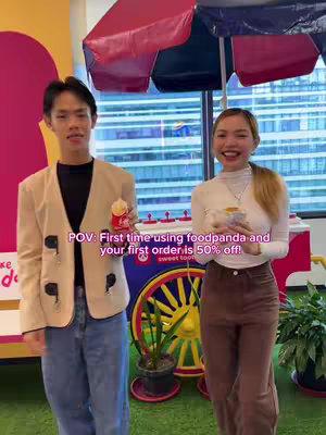 A well-lit shot featuring two people smiling while holding food items, possibly snacks. They are positioned in front of a colorful cart or stall, with a red and white striped umbrella overhead. The background includes a window view and a large pink and yellow object. The image has a positive, inviting vibe.