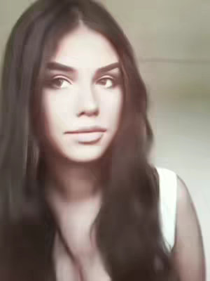 A close-up shot of a woman gazing toward the viewer. She has long, dark hair that partially frames her face, and is wearing a white top. The lighting appears soft and natural, highlighting her features.