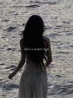 A woman stands in the water, viewed from behind, with long dark hair flowing down her back. The water is slightly rippled with a reflective surface. Overlay text reads 'she's a water sign'.
