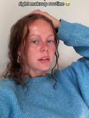 A woman with fair skin and curly brown hair is captured in a selfie-style shot. She is wearing a light blue sweater, with her right arm raised, resting her hand on her head. She has a slightly open mouth, and is looking at the camera directly, appearing to be mid-sentence. The background is a plain, off-white wall, casting a soft, diffused light on the scene.