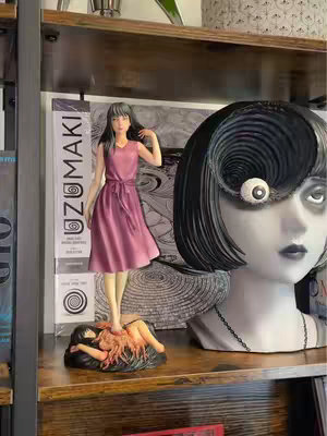 An unboxing featuring two statues on a wooden shelf. The main focus is a Tomie figure standing on a grotesque form. To the right is a large, spiral-eyed bust. The background has a book visible.