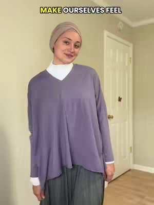 The image shows a woman with a light brown hijab, smiling at the camera. She's wearing a white turtleneck under a purple long-sleeved top and a gray skirt, standing in front of a light-colored wall with a closed white door.