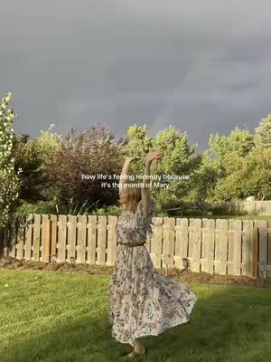 A woman in a floral dress dances with her arms outstretched in a sunlit outdoor setting, with a wooden fence and trees in the background under a cloudy sky.