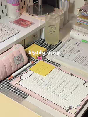 The image is a well-lit, aesthetically pleasing close-up shot of a study space. A desk is cluttered with various study-related items, including pink pencil case, pens, sticky notes, papers, a beverage in glass, and a keyboard in the background.