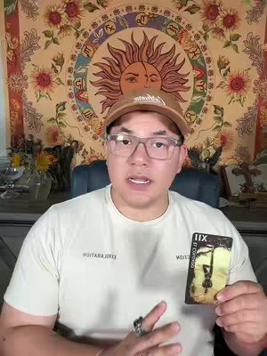 A man with glasses and a brown hat sits in front of a colorful tapestry with a tarot card in hand, speaking to the camera.