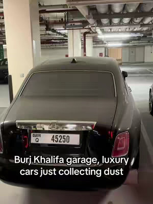 The image captures the rear of a black luxury car parked in what appears to be a parking garage. The car is covered in a thick layer of dust, signaling it hasn't been used in a while. The background shows the interior of what looks like an upscale garage, with visible infrastructure like pipes, lights, and structural supports. The car's license plate is visible, along with a hint of a red taillight. The image is composed to highlight the contrast between luxury and neglect, with the caption suggesting the car isn't being used.