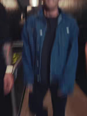 A blurry image showing a man in a denim jacket, standing in a dimly lit area. Other people are visible, and the overall effect is motion blurred and unfocused.