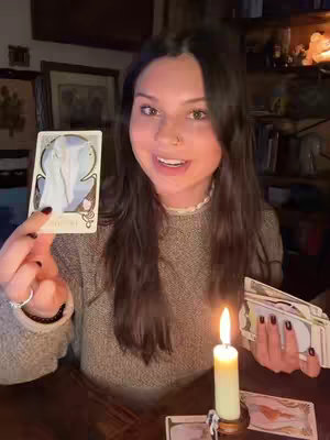 A young woman with long dark hair is holding a tarot card and a stack of cards against a background of a room with wooden furniture and bookshelves, with an illuminated candle to the right.