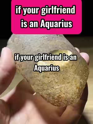 The cover image presents a hand holding a rough, light brown stone. Overlaid are two text boxes. The top box, set on a bright pink background, reads 'if your girlfriend is an Aquarius'. A second text box, positioned below closer to the stone, repeats the same phrase. The overall composition focuses on the interaction with the stone and the text