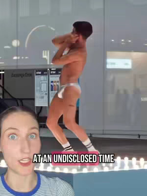A selfie-style video set against a bright, possibly indoor background of a large building. The primary focus is on a young woman with wide eyes, positioned in the foreground on the bottom left and in the center is a man dancing on what looks like a platform. The man is wearing white briefs and socks and is surrounded by the background of the building. Text is added as a hook.