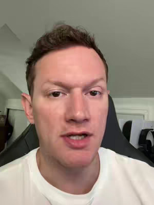 A man with short, dark hair and fair skin is positioned in a close-up shot, facing the viewer directly and speaking. He is wearing a plain white t-shirt. The background includes an out-of-focus area showing a room with a white door and some office equipment.