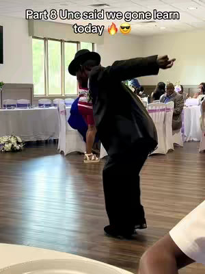 A man in a black suit and hat dances energetically on a wooden floor in a wedding reception setting. The image is a full-body shot, capturing the dynamic pose mid-dance, with a room filled with tables and guests in the background.