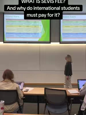 A classroom setting with two projected screens at the top of the frame displaying what appears to be tables of data. A woman stands to the right side of the frame, looking at the screens, while two students appear to be taking notes. The lighting seems to be bright and the color scheme is neutral with whites and hints of green.