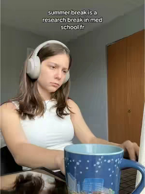 A young woman wearing headphones sits in front of a laptop with a blue mug on the desk. Text overlayed states summer break is a research break in med school fr.