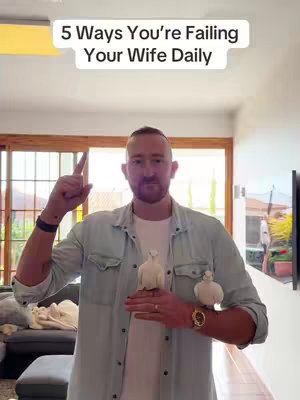 A man with a beard and short hair stands indoors, holding two white bird figurines in front of him. He is wearing a light blue chambray shirt over a white t-shirt, with a gold watch on his wrist. The background includes a wooden door and window, with a glimpse of the outside. Text overlay at the top reads '5 Ways You're Failing Your Wife Daily'.