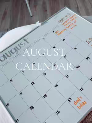 A calendar is displayed at an angle against a blurred background, likely of a wooden floor. The calendar is white, with the month of August highlighted in large, silver letters at the top. The phrase "AUGUST CALENDAR" is written in white letters.