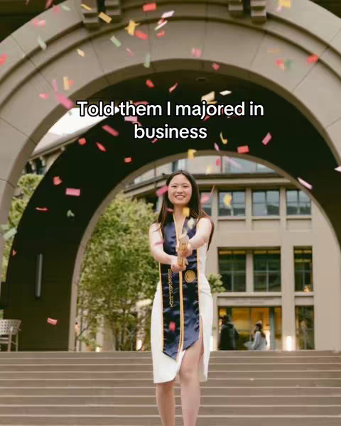 A young woman in a white dress and graduation sash stands in front of a large stone archway. She is smiling and holding what appears to be a confetti cannon. Confetti is falling around her. The background includes a building with large windows and a grey sky.