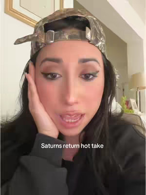 The image shows a close-up shot of a woman speaking, with a camo baseball cap on her head. The text overlay says 'Saturns return hot take'. Her hand is raised to her cheek, and she appears to be in an indoor setting.