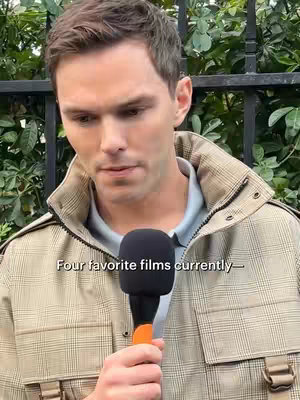 A medium shot shows a man holding a microphone, with a city fence and greenery behind him. The man is fair-skinned with short brown hair, wearing a tan jacket over a light blue collared shirt and looking slightly off-camera, with text overlayed at the bottom.