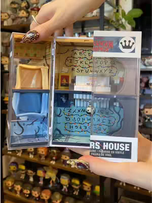 A person's hands presenting a small display case with a Stranger Things theme, filled with miniature rooms and characters. The case is layered, with the person's hand in the foreground showcasing a manicure with decorative nails and a ring. The background has shelves with many figurines. The overall feel is detailed, colorful, and designed to appeal to fans of the show.