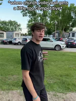 A young man is standing in a grassy area, looking slightly off-camera with a surprised expression. The background shows a few cars, a row of buildings, and trees under a partly cloudy sky.