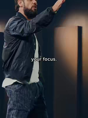 A man dressed in a black jacket, white t-shirt, and blue jeans is gesturing upwards with his right hand. The text 'your focus' is overlaid on the image in a white font.