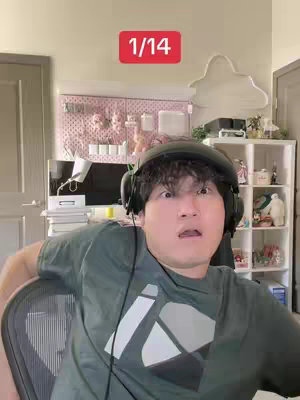 A person with dark hair wearing a headset and a green t-shirt is shown indoors. They have a surprised expression with wide eyes and an open mouth.  Behind him is a wall with a white cloud-shaped object, a pink grid/pegboard, and shelves. The image is captioned with the number 1/14 in a red box at the top of the image.