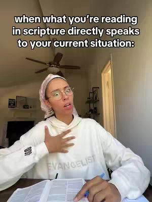 A person with glasses, a headscarf, and a white sweatshirt is looking up in surprise with their hand on their chest. In front of them is an open book and a pen.