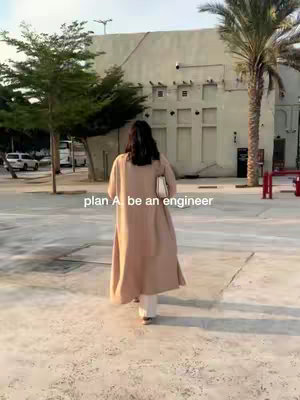 A woman with long dark hair walks away from the camera on a paved area, buildings and greenery are in the background, with added text.