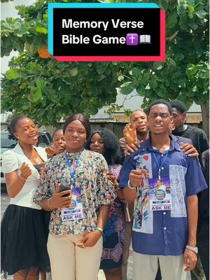 The image shows a group of people outdoors, possibly participating in a game or event. There is a text overlay at the top indicating that this is a "Memory Verse Bible Game". The group is diverse, with people of different genders and skin tones. They are gathered in front of a blurred background that appears to be a tree or other greenery.