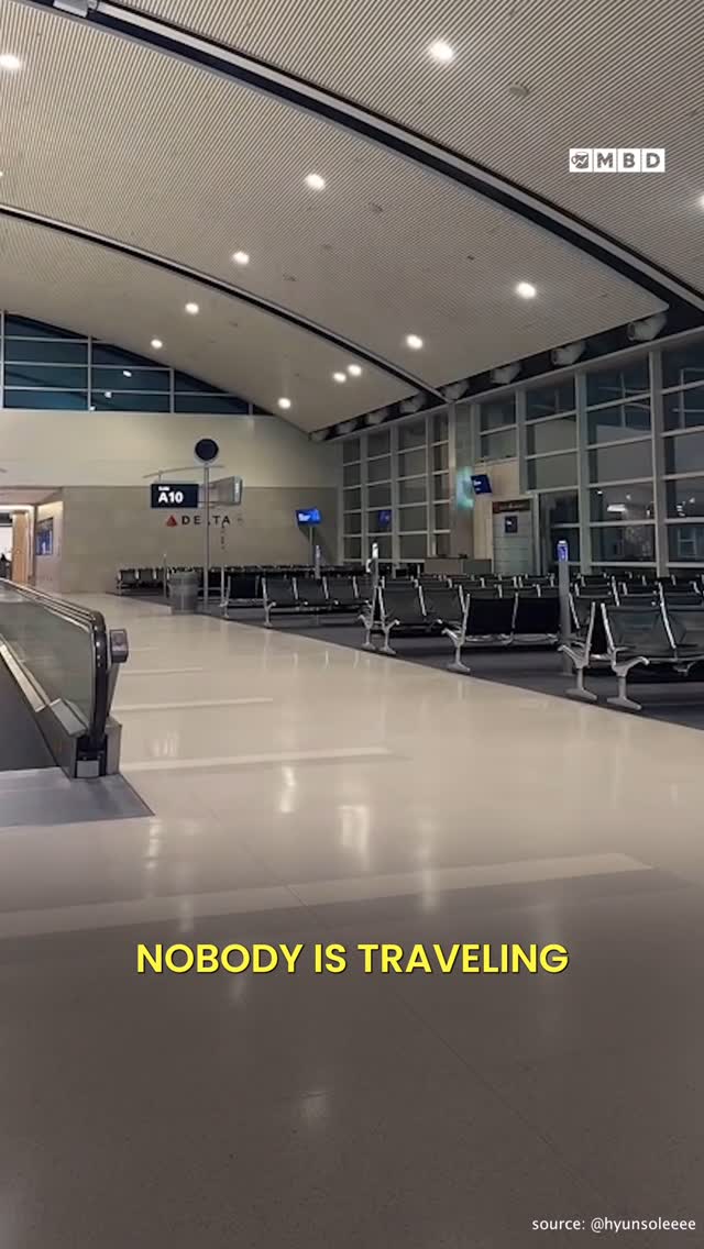An empty airport terminal with rows of seats and an automated walkway. The ceiling is high with recessed lighting, and large windows provide a view of the outside. A yellow text overlay at the bottom reads: NOBODY IS TRAVELING