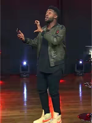 A man in a bomber jacket and sneakers is speaking with his hands raised, possibly preaching or giving a speech, on a stage. The background features stage lighting and a dark backdrop.