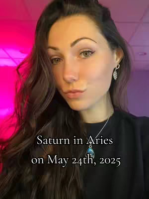 A young woman with long brown wavy hair is centered in the frame, looking slightly towards the camera. The background has a gradient of bright pink and purple. Text overlayed on the image reads 'Saturn in Aries on May 24th, 2025'. She is wearing a black top and a silver necklace with a pendant. The overall composition is a selfie with a close-up focus.