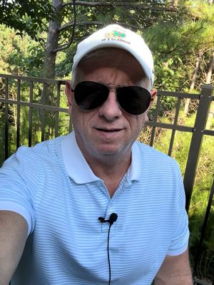 A middle-aged man with short, light-colored hair and sunglasses is seated outside, possibly on a patio. He wears a light blue polo shirt and a white cap with a logo. A microphone is clipped to his collar. Behind him, a dark metal fence and trees with green foliage are visible, suggesting a natural outdoor setting.