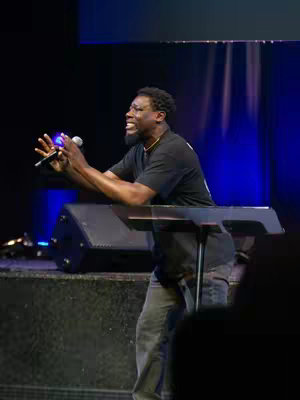 An image of a Black man speaking into a microphone at a podium in front of a dark stage with blue ambient lighting. He is wearing a black t-shirt, gray jeans, and has his hair styled in short curls. He appears to be mid-sentence, with an open mouth and raised hands. A dark stage backdrop is partially visible behind him.