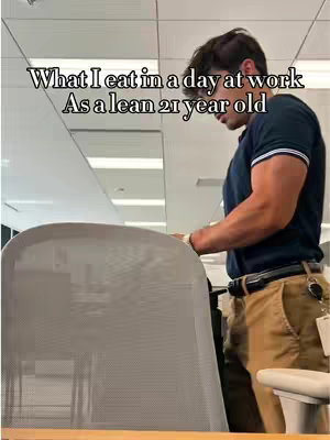 A man leans against an office chair in an office setting. The man is facing to the side, wearing a dark shirt and tan pants. Text overlays appear at the top, detailing content about what he eats in a day.