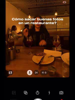The cover image shows a woman sitting at a restaurant table, seemingly filming a video. The setting is dim, with warm lighting from hanging lights. A glass of red wine, plates with food remnants, and other dining elements are visible. Text overlay reads "Cómo sacar buenas fotos en un restaurante?" (How to take good photos in a restaurant?). The scene appears to be captured using the phone camera in front of her, with its menu and settings visible at the bottom of the screen.