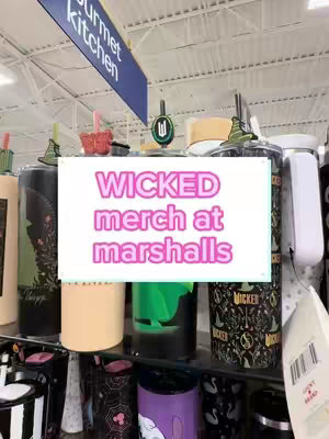 A photo showcasing merchandise from the musical 'Wicked' at Marshalls. The image is filled with various products, including tumblers and other items, displayed on store shelves. A white rectangular overlay prominently displays the text 'WICKED merch at marshalls', highlighted by a pink drop shadow. The background shows the store's ceiling and signs. The overall composition utilizes a medium shot with a focus on the merchandise display.