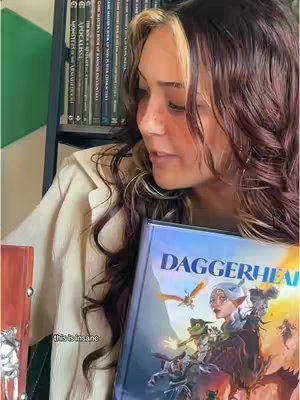 A woman with lightened hair holds a book titled 'Daggerheart' in front of her. She is in a room with bookshelves.