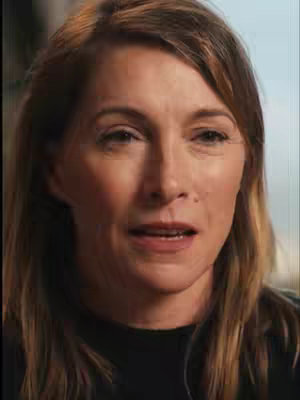 A medium shot of a woman with light brown hair, looking slightly to the side with a neutral expression. The background is blurred.