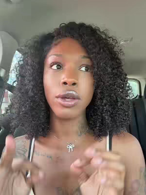 A woman with curly black hair is positioned inside a car, looking towards the side, as if talking. She is wearing a black and white spaghetti strap top and a delicate bow necklace. The car's interior, including the headrest, is visible. The background suggests an outdoor setting obscured by the car.