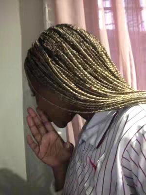 A close-up shot of a person's head and shoulder, their face partially obscured. They have braided hair, which is primarily a golden brown color. The individual is wearing a white and red striped shirt that is unbuttoned. Their hand is up to their face, as if they are whispering or shielding something from view.